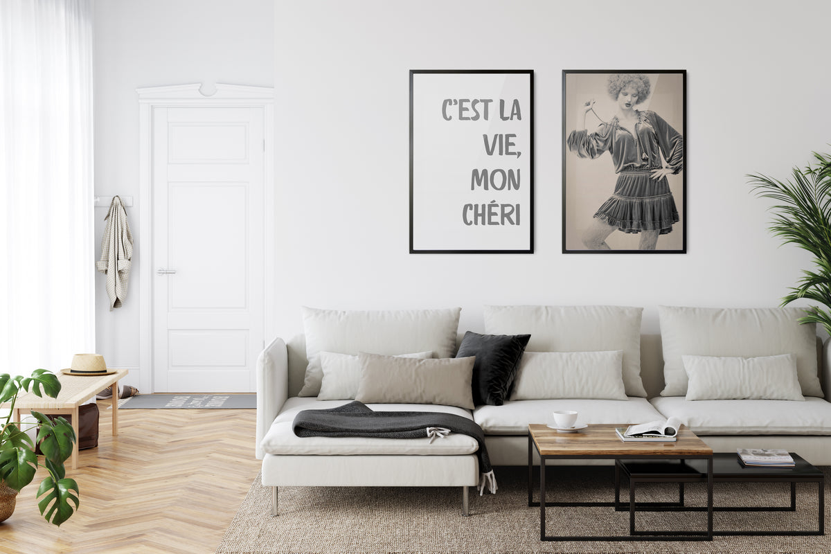 a living room setting with two framed images on the wall. One is a poster with the words C'est La Vie, Mon Cheri. The other is a vintage looking woman with a curly wig
